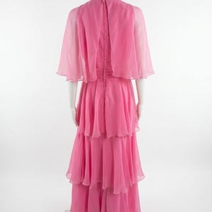 May include: A pink, tiered formal gown with a high neckline and a sheer cape. The dress is displayed on a mannequin, showcasing its flowing, layered skirt and delicate fabric. The back of the dress features a zipper closure.