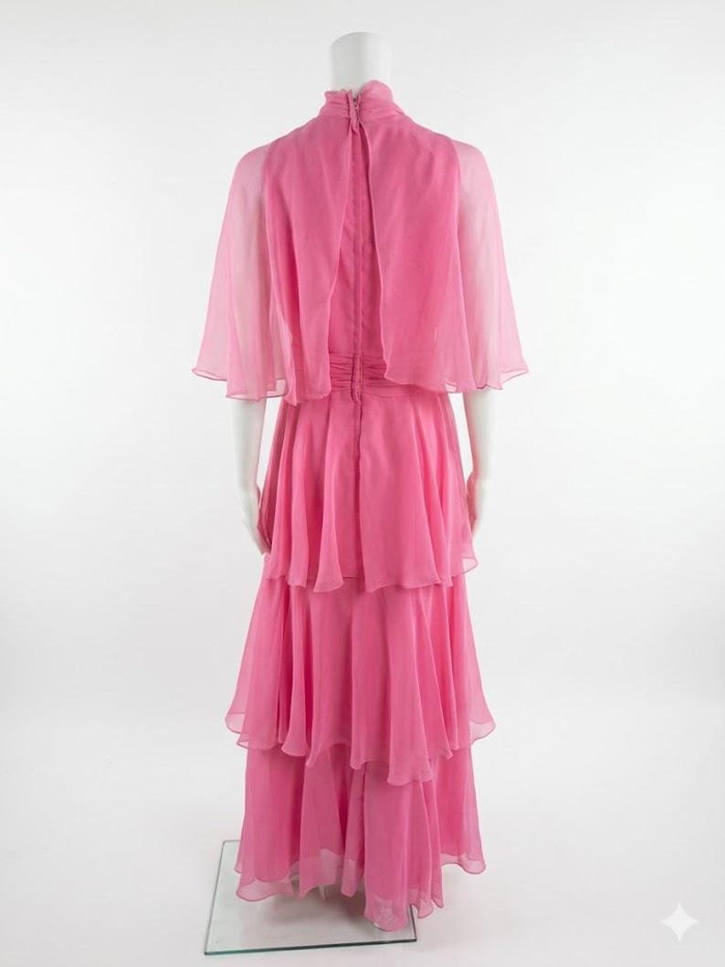 May include: A pink, tiered formal gown with a high neckline and a sheer cape. The dress is displayed on a mannequin, showcasing its flowing, layered skirt and delicate fabric. The back of the dress features a zipper closure.