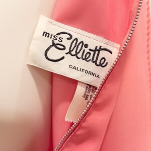 May include: Close-up of a pink garment with a zipper. A white label with the text "miss Elliotte CALIFORNIA" is sewn into the garment. The label has black cursive text.