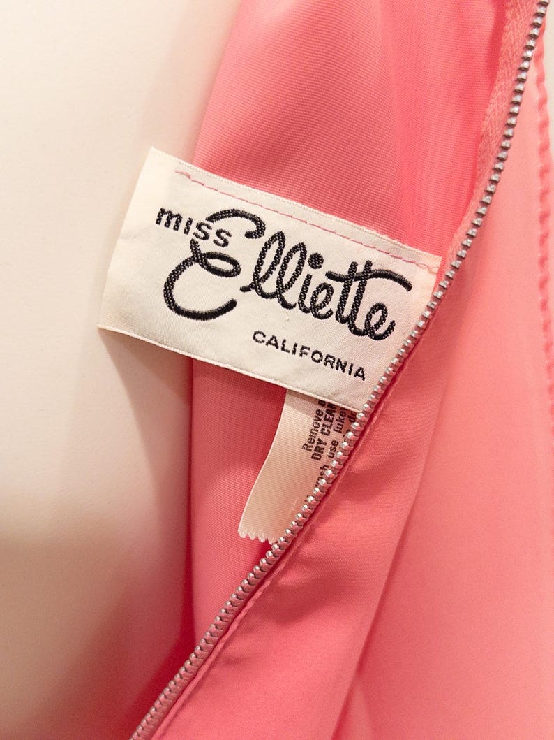 May include: Close-up of a pink garment with a zipper. A white label with the text "miss Elliotte CALIFORNIA" is sewn into the garment. The label has black cursive text.