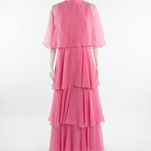 May include: A bright pink, tiered maxi dress with a matching cape. The dress features a high neckline and a flowing, layered skirt. The cape is sheer and drapes over the shoulders. The dress is displayed on a mannequin.
