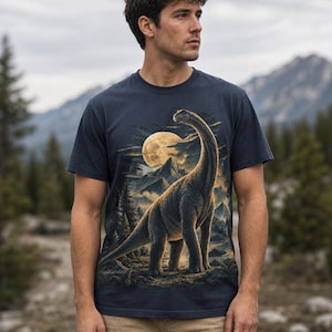 Könnte beinhalten: Ein dunkelblaues kurzärmeliges T-Shirt mit einem Grafikdruck eines langhalsigen Dinosauriers in einer Landschaft mit Kiefern, Bergen und einem großen Vollmond im Hintergrund. Das Motiv verwendet Gold-, Braun- und Dunkelblautöne.