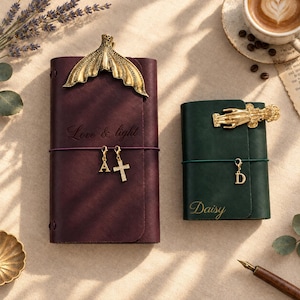 May include: A dark purple leather journal with a gold mermaid tail clip, engraved with "Love & light" and two charms, "A" and a cross. Beside it, a dark green leather journal with a gold hand clip, engraved with "Daisy" and a "D" charm.