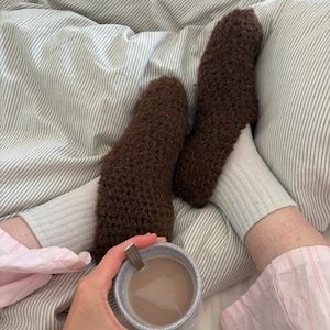 May include: A pair of dark brown fuzzy crochet slippers worn over light grey ribbed socks. A ceramic mug with a light brown beverage and a metal spoon is held in the foreground. The slippers feature a textured, looped yarn construction.