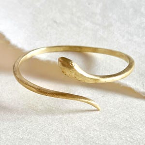 May include: A gold-colored snake-shaped bracelet. The bracelet wraps around in a spiral, with the snake's head at one end and a pointed tail at the other. The snake's head has a small, round detail representing an eye.