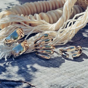 May include: Four gold-colored rings with unique designs are displayed on a weathered wooden surface. One ring has a curved, open design, while another features a spiral shape. A third ring has a rounded shape, and the fourth ring has a rectangular shape.
