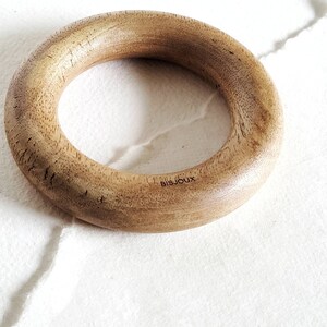 May include: A wooden ring with a smooth, light brown finish. The ring has a slightly textured surface and the word "BISJOUX" is engraved on the outside.