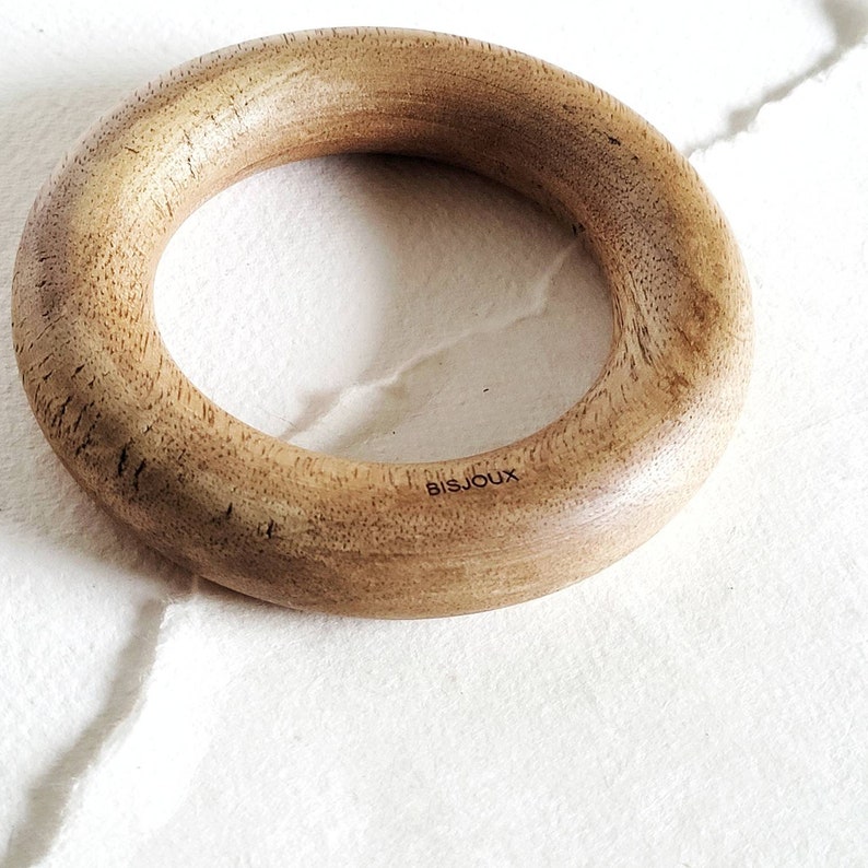 May include: A wooden ring with a smooth, light brown finish. The ring has a slightly textured surface and the word "BISJOUX" is engraved on the outside.