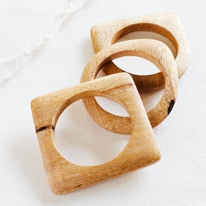 May include: Three wooden bangles, each with a different shape. The bangles are stacked on top of each other, with the largest bangle at the bottom and the smallest bangle at the top. The bangles are all made of a light brown wood.