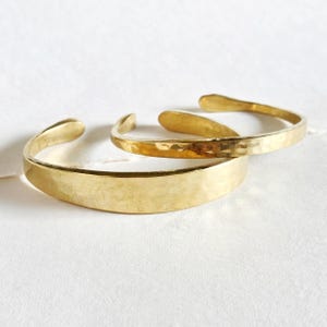 May include: Two gold-toned cuff bracelets with a hammered, textured finish. The bracelets are open-ended, featuring a polished metallic surface that reflects light, showcasing a simple, curved design with rounded ends for a smooth appearance.