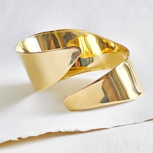 May include: A gold-toned metal cuff bracelet with a unique, twisted design. The bracelet is smooth and polished, with a modern and minimalist aesthetic.
