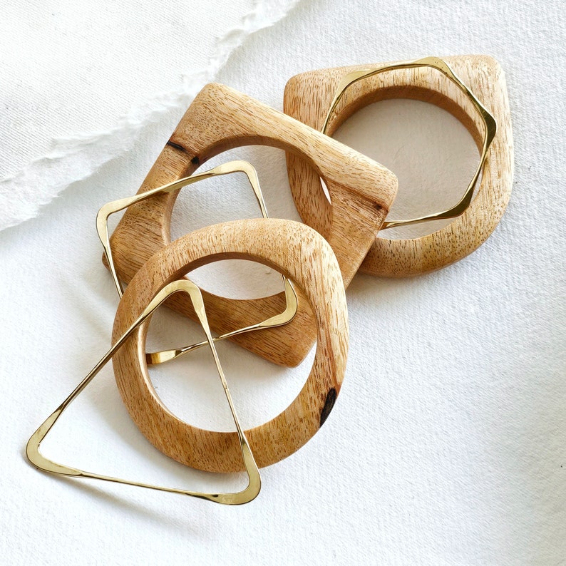 May include: Three wooden bangles with gold metal accents. The bangles are in the shape of a square, a circle, and a triangle.