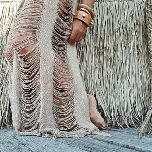 May include: A beige crochet skirt with a distressed, frayed design. The skirt is worn by a person with a wooden bangle bracelet on their wrist.