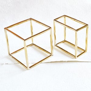 Può includere: Due forme di cubo geometrico in filo metallico dorato. Il cubo più grande è una forma tridimensionale, mentre il cubo più piccolo è una forma bidimensionale.