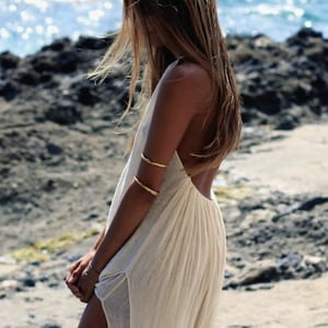 May include: A sleeveless, off-white, floor-length dress with a deep back and side slits. The garment is accessorized with two thin, polished gold-tone metal arm cuffs worn on the upper arm, creating a simple and clean aesthetic for the ensemble.