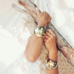 May include: Two gold-toned metal bangles, one is a large, round, dome-shaped bangle, the other is a smaller, curved bangle. The bangles are worn on the wrists of a person.