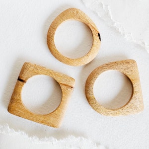 May include: Three wooden bangles in different shapes: a teardrop, a square, and a rounded rectangle. The bangles are all made of light brown wood with a natural finish.