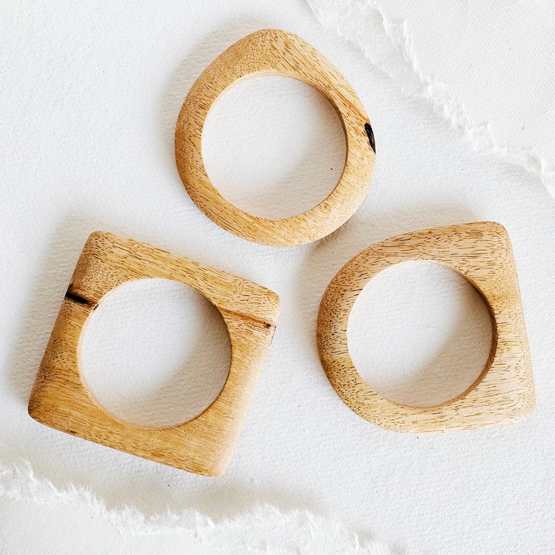 May include: Three wooden bangles in different shapes: a teardrop, a square, and a rounded rectangle. The bangles are all made of light brown wood with a natural finish.