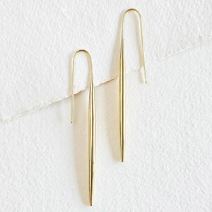 May include: A pair of gold-toned earrings with a long, thin, pointed design. The earrings are made of a smooth metal and have a simple, minimalist style.