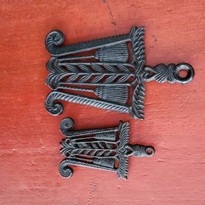 Może przedstawiać: Dwa czarne żeliwne podstawki z dekoracyjnym wzorem kłosów pszenicy i pętlą. Podstawki znajdują się na czerwonym drewnianym podłożu.