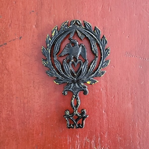 May include: Black cast iron trivet with a design of a bird with a heart in a wreath. The trivet has a yellow patina.