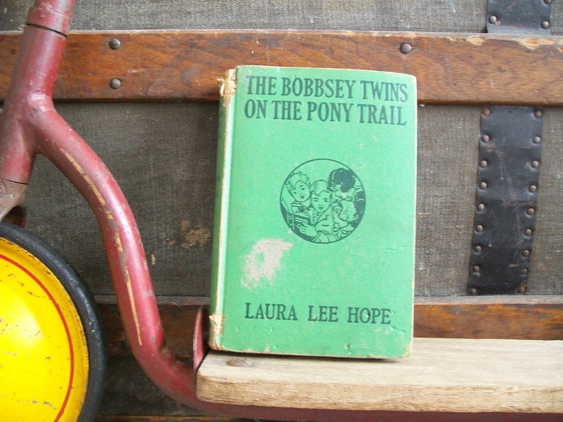 May include: A vintage green book titled "The Bobbsey Twins on the Pony Trail" by Laura Lee Hope. The book is sitting on a wooden shelf.