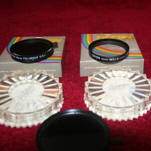 May include: Two Tiffen brand camera lens filters in their original packaging. The filters are black and round. The packaging is white with rainbow stripes and black text. The text on the packaging reads "Tiffen 43mm Sky 1-A U.S.A." and "Tiffen 43mm Polarizer U.S.A."