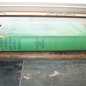 May include: A green hardback book titled "The Twin's On The Pony Trail" by Laura Lee Hope. The book is published by Grosset & Dunlap.