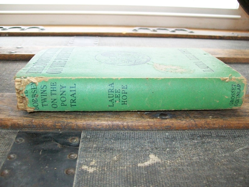 May include: A green hardback book titled "The Twin's On The Pony Trail" by Laura Lee Hope. The book is published by Grosset & Dunlap.