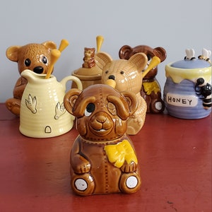 May include: A collection of five ceramic honey bear jars. The jars are decorated with brown, yellow, and blue paint. One jar has the word "Honey" written on it. The jars are sitting on a red surface.