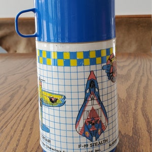 Puede incluir: Termo azul y amarillo con un patrón de cuadrícula e imágenes de aviones. El termo tiene una tapa azul con un asa. El texto en el termo dice "F-19 STEALTH ALADDIN'S Characters From Cleveland, Inc."