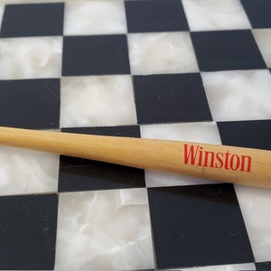 Könnte beinhalten: Ein Miniatur-Baseballschläger-Stift aus Holz mit einem roten "Winston"-Logo auf einer schwarz-weißen Schachbrettfläche.