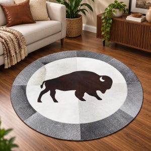 Pode incluir: Um tapete circular com a silhueta castanho-escura de um bisão centrada num fundo branco. O tapete é contornado por um anel largo e segmentado de retalhos alternados em cinzento-claro e cinzento-escuro, criando um padrão geométrico.