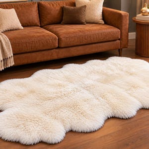 May include: An off-white, long-pile sheepskin rug with an irregular, organic shape. The rug features a dense, fluffy texture and is laid flat on a wooden floor, showcasing its natural, curved edges and soft, cream-colored fibers.
