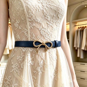 May include: A navy blue belt with a gold-toned metal bow-shaped buckle worn over a sleeveless cream-coloured dress featuring an intricate floral lace overlay pattern that extends from the bodice down to the skirt of the garment.