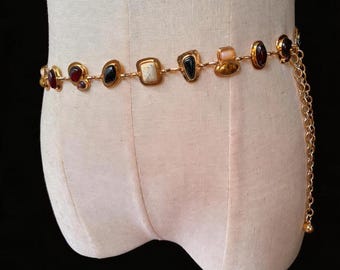 May include: A gold-tone chain belt featuring a series of alternating geometric links and smooth, polished stones in shades of deep red, black, cream, and amber. The belt includes a dangling chain section on one side with a small spherical end piece.