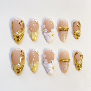May include: A set of ten almond-shaped press-on nails featuring a nude base. Designs include gold metallic swirls with small white pearls, pale yellow textured waves, white 3D floral accents, and white lace patterns with gold beaded horizontal lines.