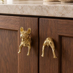 May include: Two gold-coloured metal cabinet knobs shaped like a French Bulldog. One knob depicts the front view of the dog with ears, face, and front legs, while the other knob depicts the rear view of the dog with hind legs and a tail.
