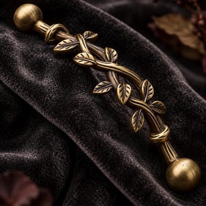 May include: A brass-toned metal cabinet handle featuring a central vine design with detailed leaves. The handle has a rounded ball-shaped finial at each end and a curved, textured body that mimics the appearance of intertwined branches.