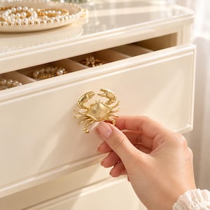 Peut inclure: Une poignée de tiroir en métal doré en forme de crabe fixée sur un tiroir de commode en bois crème. Le tiroir est entrouvert, révélant des compartiments intérieurs contenant divers bracelets de perles et des bijoux.