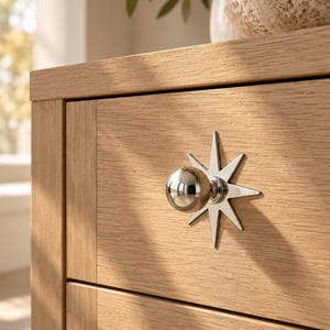 May include: A light-colored wooden drawer front featuring a polished silver-toned metal knob. The knob is centered on a decorative starburst-shaped backplate with eight pointed rays, creating a reflective metallic accent on the wood grain.