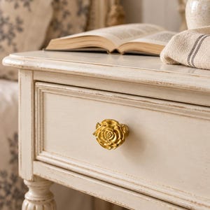 Könnte beinhalten: Ein goldfarbener Schubladenknopf in Form einer blühenden Rose, befestigt an der Vorderseite eines cremefarbenen Nachttischs aus Holz mit abgenutzter Oberfläche. Der Knopf zeigt detaillierte, geschichtete Blütenblätter.