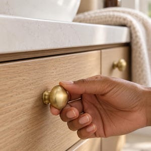 May include: A round, brushed brass cabinet knob with a smooth, metallic finish attached to a light-toned, horizontal wood grain drawer front. The knob features a simple, circular design that protrudes from the flat surface of the drawer.
