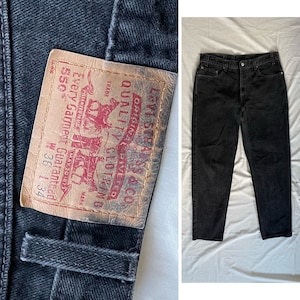 Pode incluir: Um par de calças de ganga pretas Levi Strauss & Co. com um remendo de couro castanho que diz "Original Riveted Quality Clothing" e "Every Garment Guaranteed 550" com o tamanho W38 L34.