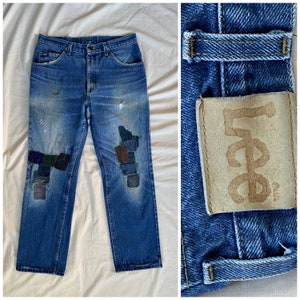 Op de afbeelding: Blauwe denim jeans met meerdere patches die op de knieën zijn genaaid. De jeans hebben een verbleekte look en een bruin lederen Lee-label op de achterzak.
