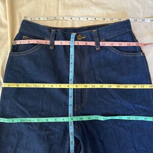 K&ouml;nnte beinhalten: Dunkelblaue Jeans mit Ma&szlig;en, die mit Ma&szlig;b&auml;ndern angezeigt werden. Die Jeans hat einen Knopfverschluss und Taschen. Taille und Schrittl&auml;nge werden mit rosa, gelben und blauen Ma&szlig;b&auml;ndern gemessen. Die Jeans liegt flach auf einer beigen Oberfl&auml;che.
