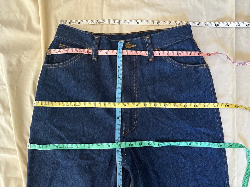 K&ouml;nnte beinhalten: Dunkelblaue Jeans mit Ma&szlig;en, die mit Ma&szlig;b&auml;ndern angezeigt werden. Die Jeans hat einen Knopfverschluss und Taschen. Taille und Schrittl&auml;nge werden mit rosa, gelben und blauen Ma&szlig;b&auml;ndern gemessen. Die Jeans liegt flach auf einer beigen Oberfl&auml;che.
