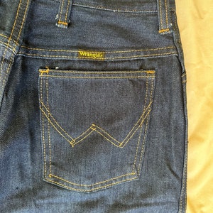 K&ouml;nnte beinhalten: Nahaufnahme einer dunkelblauen Jeans mit einer Ges&auml;&szlig;tasche mit einem markanten 'W'-Nahtdesign. Der Markenname 'Wrangler' ist auf dem Bund sichtbar. Die Jeans hat gelbe N&auml;hte und einen klassischen Vintage-Stil.