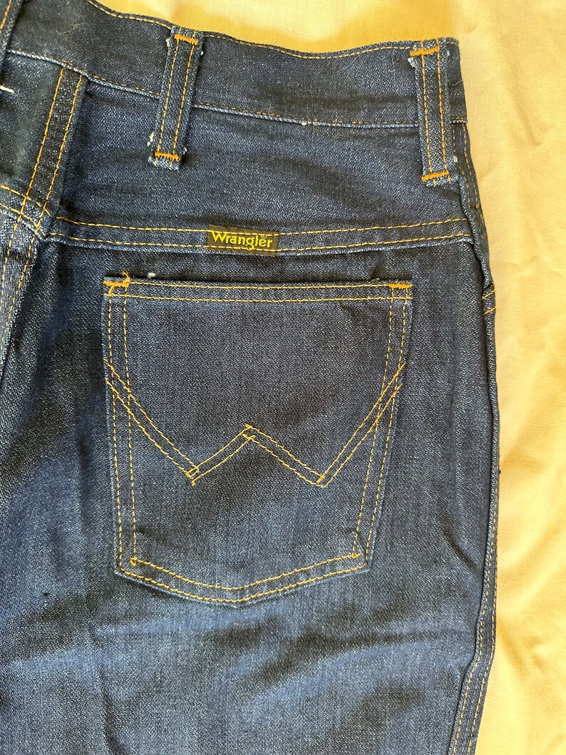 K&ouml;nnte beinhalten: Nahaufnahme einer dunkelblauen Jeans mit einer Ges&auml;&szlig;tasche mit einem markanten 'W'-Nahtdesign. Der Markenname 'Wrangler' ist auf dem Bund sichtbar. Die Jeans hat gelbe N&auml;hte und einen klassischen Vintage-Stil.