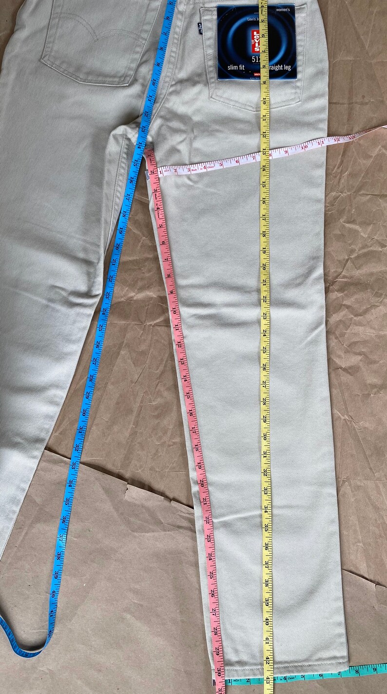 May include: A pair of light beige jeans with a Levi's 511 slim fit straight leg label on the back pocket. The jeans are measured with three tape measures, one blue, one pink, and one yellow.